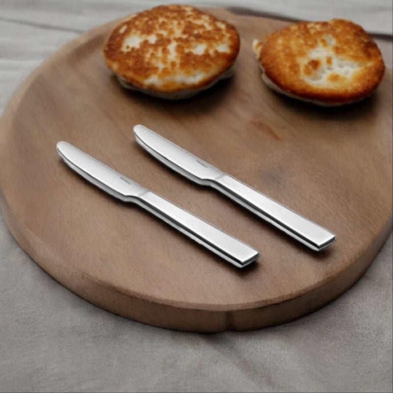 Two polished stainless steel Trendz dinner knives with flat ergonomic handles on round wooden board beside two golden-brown baked items.