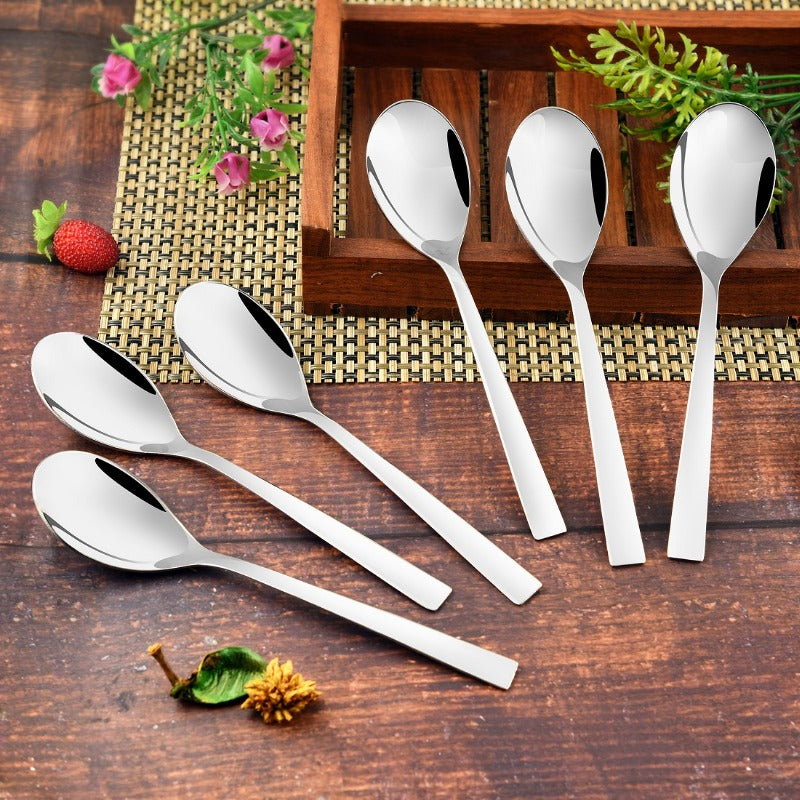Six stainless steel teaspoons with flat modern handles arranged on rustic wooden surface with bamboo mat, pink flowers, strawberry, and decorative elements, three spoons resting on dark wooden holder