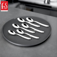Six mirror-polished stainless steel teaspoons with curved handles arranged in three rows on round dark gray plate, FnS Premium Dining logo visible