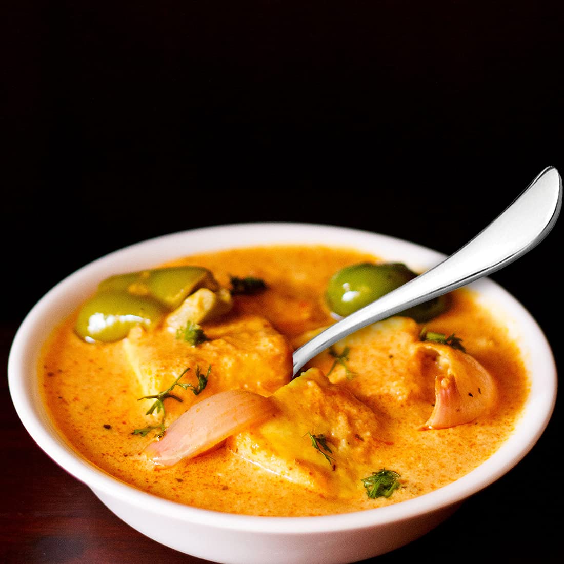 FNS Victoria serving spoon in white bowl filled with curry dish against dramatic black background, showcasing mirror finish and elegant curved handle design.