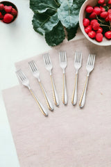 Six silver dinner forks with gold accent handles arranged on beige napkin with strawberries