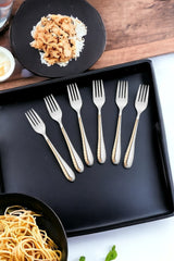 Six silver forks with gold detailed handles displayed on black serving tray with pasta