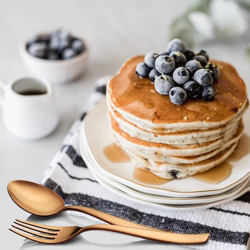 Stack of blueberry pancakes with syrup, paired with elegant rose gold fork and spoon on table.