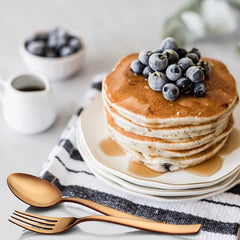 Stack of blueberry pancakes with syrup, paired with elegant rose gold fork and spoon on table.