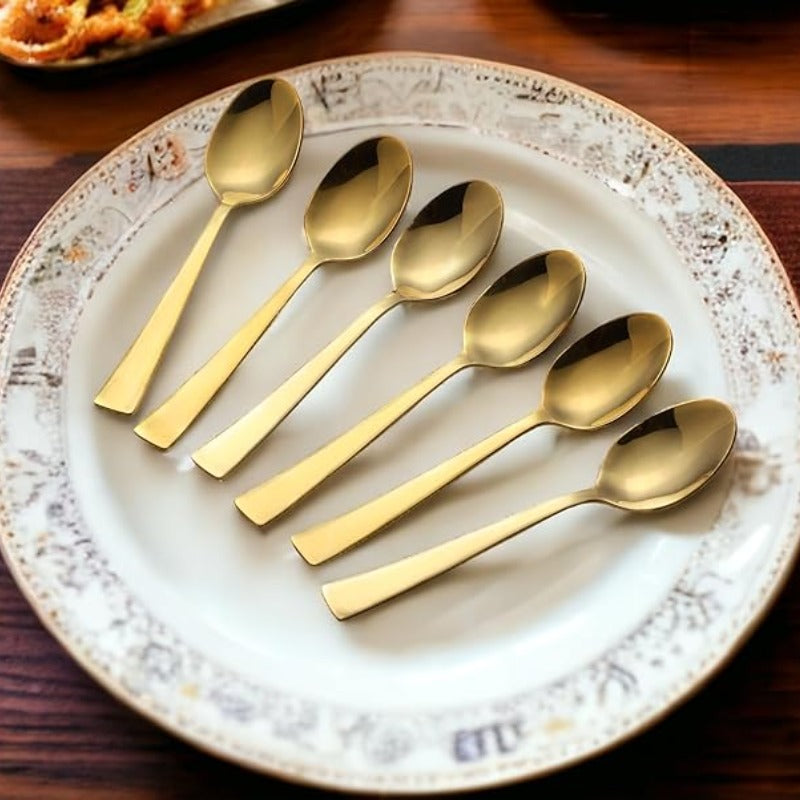 Six gold dessert spoons with flat handles arranged on vintage white decorative plate on wooden table lifestyle setting
