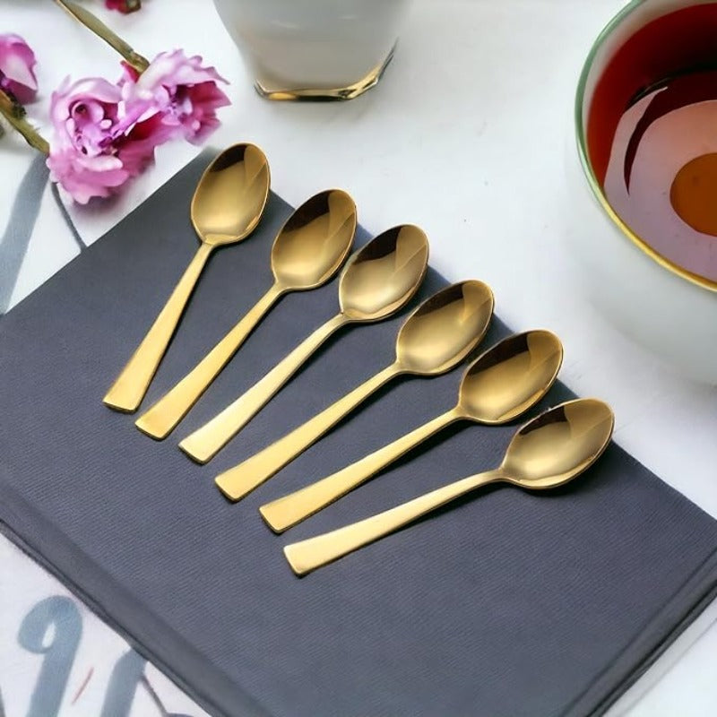 Six gold tea spoons arranged on grey napkin with pink flowers and tea cup in elegant lifestyle setting