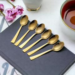 Six gold tea spoons arranged on grey napkin with pink flowers and tea cup in elegant lifestyle setting