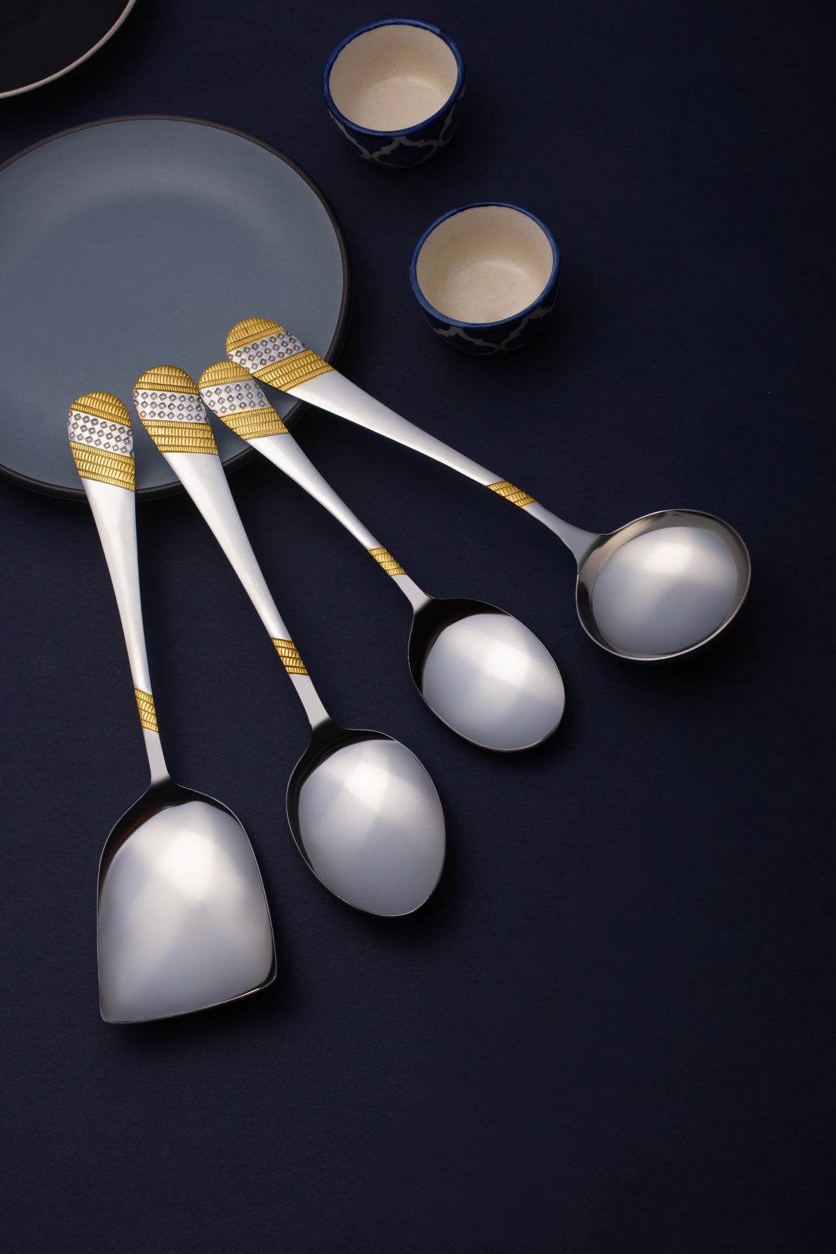 Four stainless steel serving utensils with gold and silver handles displayed on navy blue surface beside gray plate and decorative blue bowls.