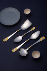 Four-piece serving spoon set with decorative gold handles artistically arranged on dark navy background with gray plate and blue ceramic bowls.