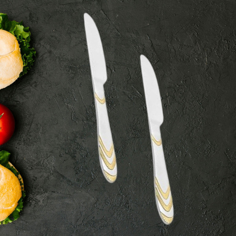 Two Magna stainless steel dinner knives with gold wave pattern handles on dark textured surface beside fresh lettuce, tomato, and burger buns.