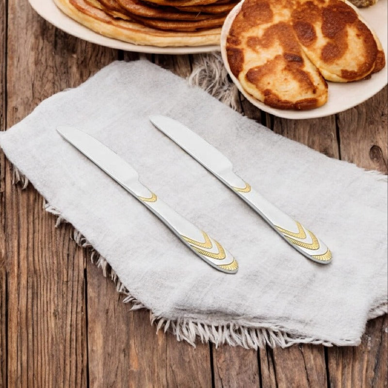 Three Magna dinner knives with elegant gold chevron pattern handles on white napkin beside pancake plates on rustic wooden table.