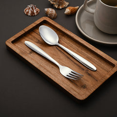 Polished spoon and fork on wooden tray, styled with seashells and coffee cup on dark background.
