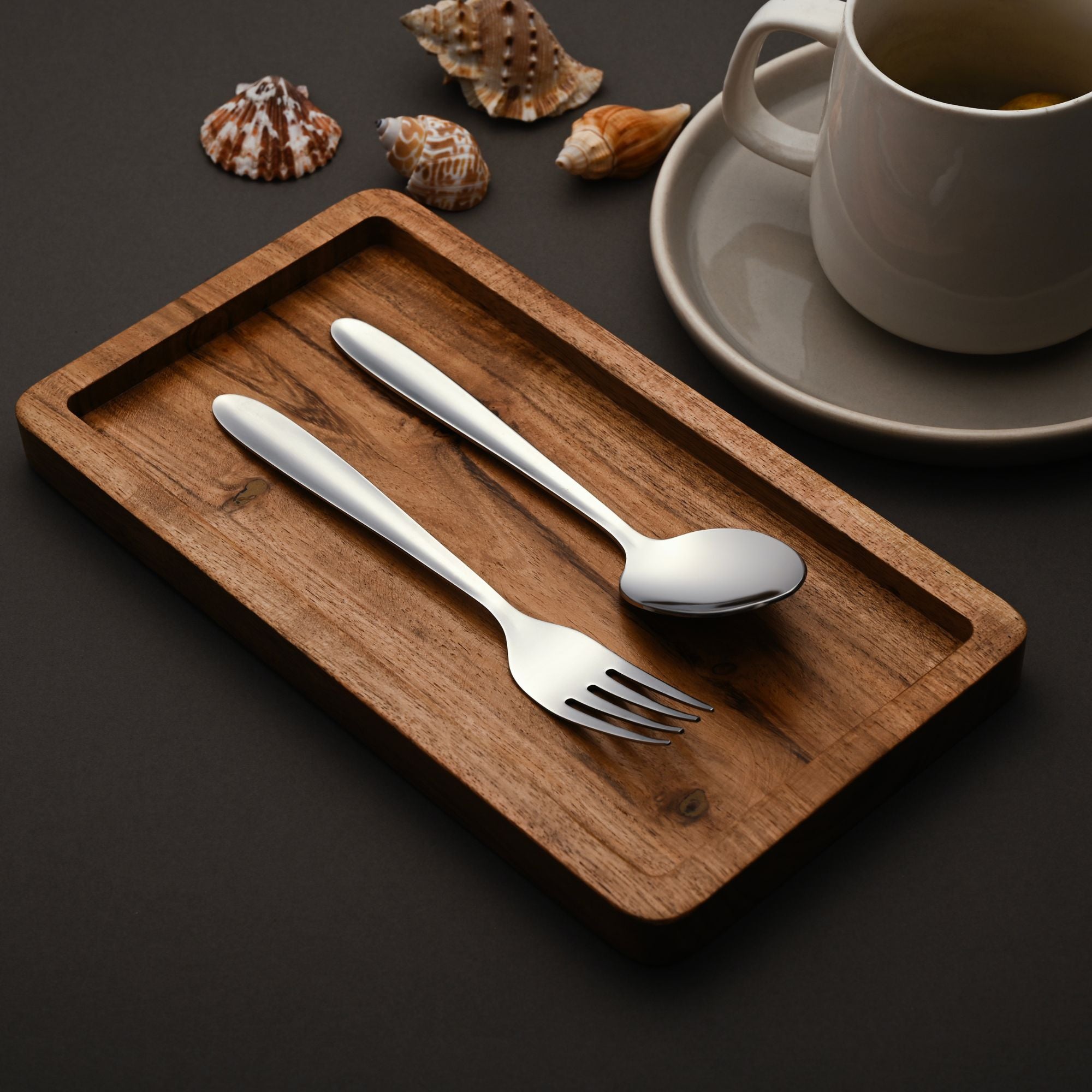 Stainless steel spoon and fork on wooden tray, beside a tea cup and decorative seashells.