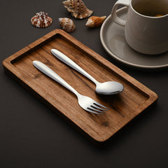 Stainless steel spoon and fork on wooden tray, beside a tea cup and decorative seashells.