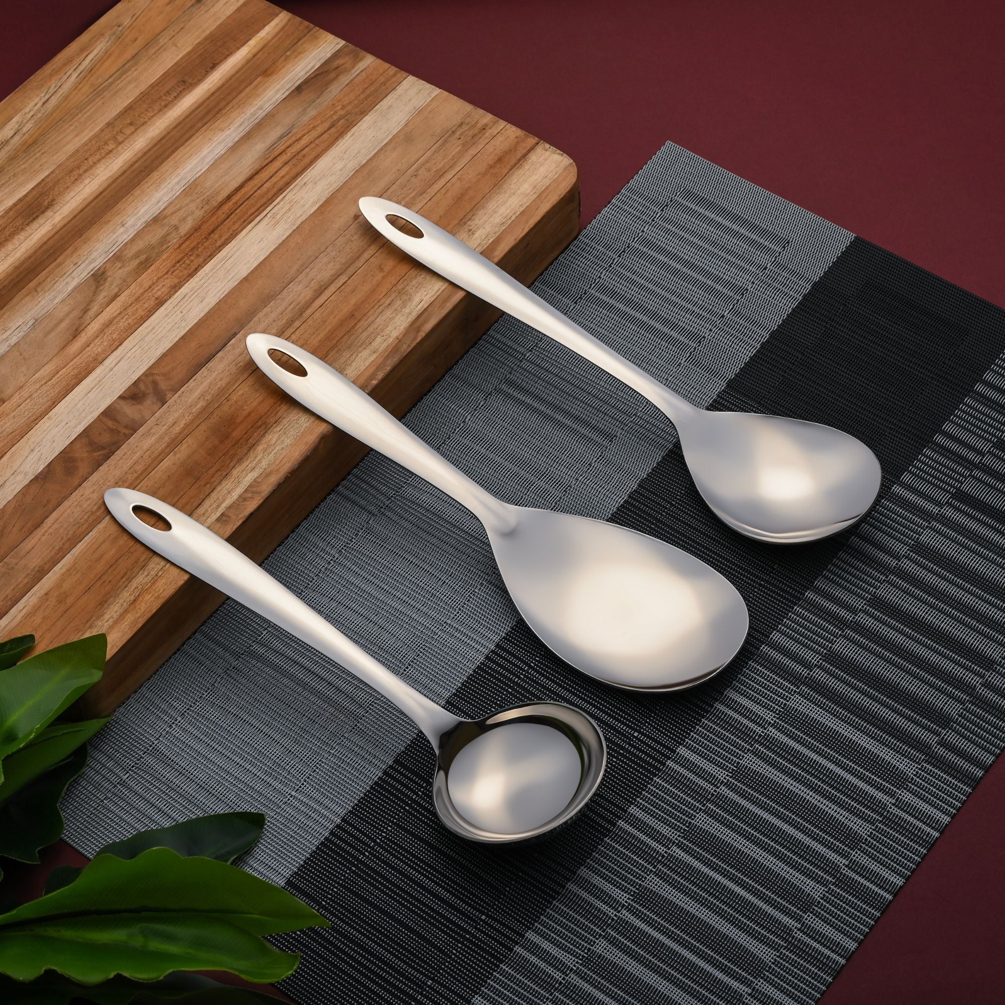 Montavo by FNS Amazon 3-piece stainless steel serving set overhead view on striped wooden cutting board with gray textured placemat and green plant