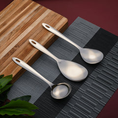 Montavo by FNS Amazon 3-piece stainless steel serving set overhead view on striped wooden cutting board with gray textured placemat and green plant