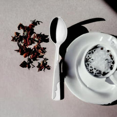 White teaspoon beside coffee cup on saucer with dried hibiscus petals, creating artistic shadow on light surface in overhead view.