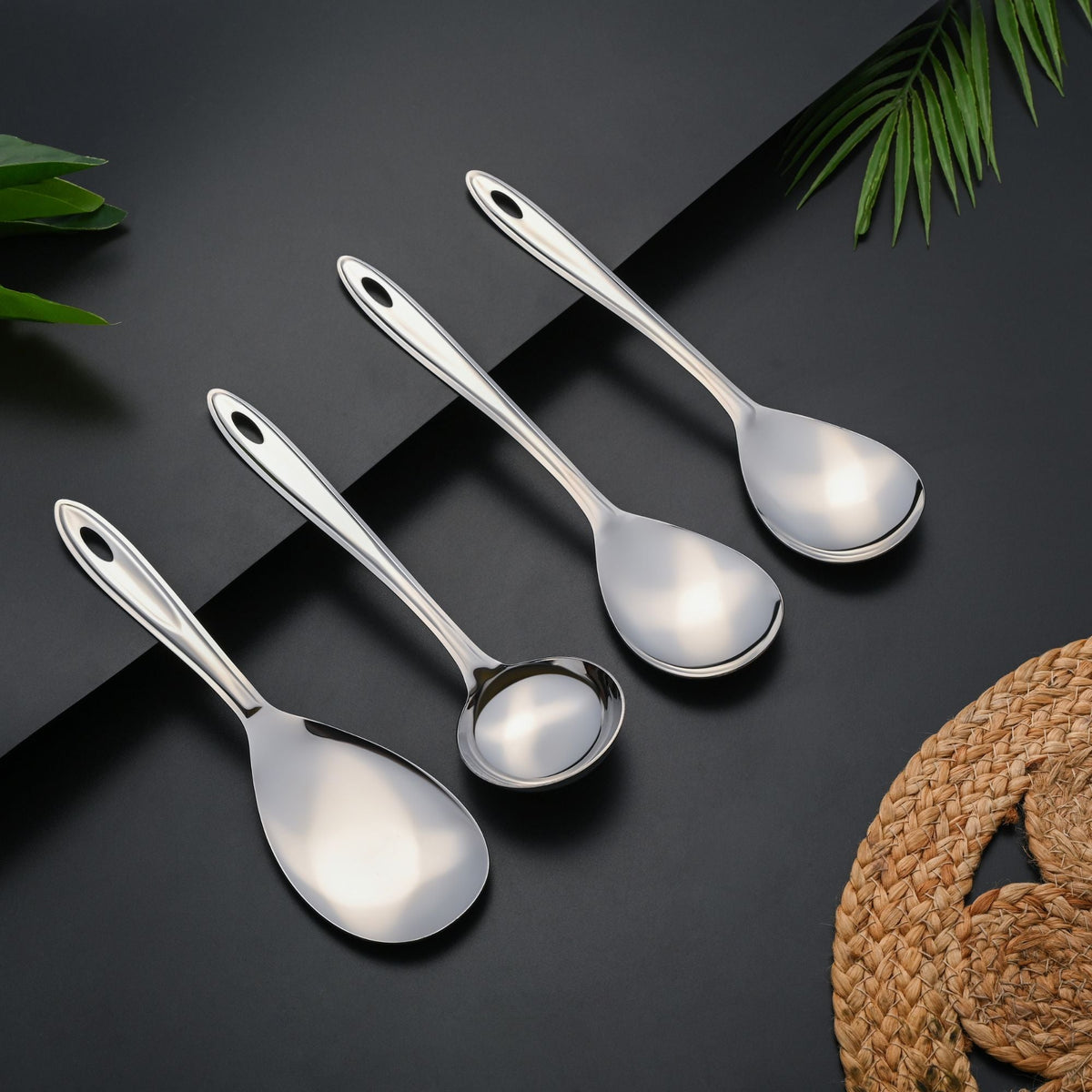 Trinity serving spoons displayed on grey and black background with green plant leaves, woven basket decoration, and elegant shadow effects.