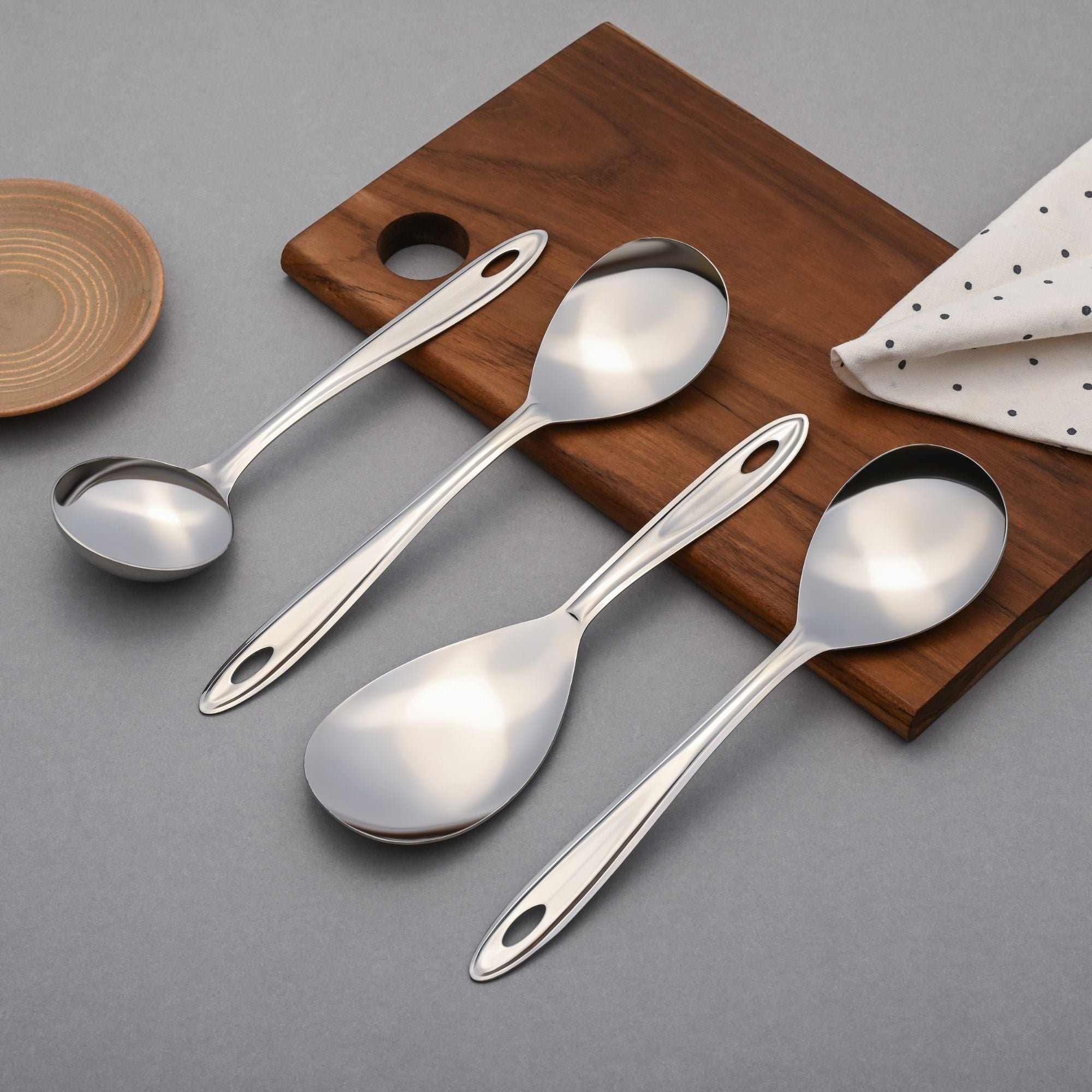 Trinity serving spoons set displayed on wooden holder with polka dot napkin, circular wooden plate, and grey background styling.