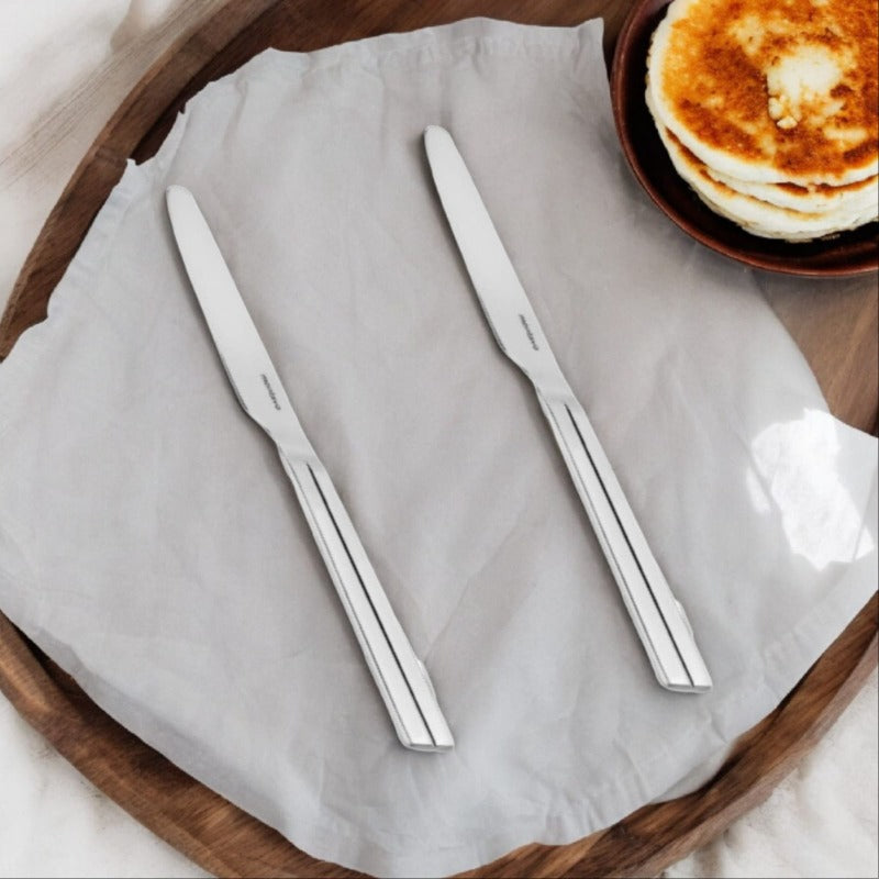 Two polished stainless steel Nile dinner knives with sleek linear handles on white napkin beside wooden tray with stack of pancakes.