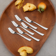Six stainless steel dinner forks with textured handles neatly arranged on a large wooden plate, alongside partially eaten fruit and cake pieces.