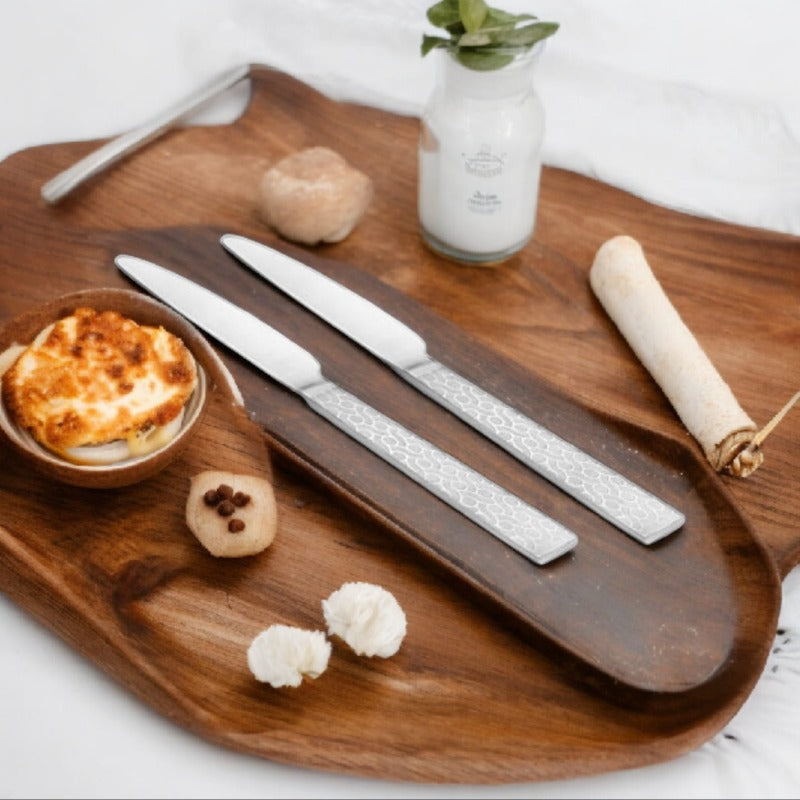 Styled flat lay of elegant dining setup featuring two silver textured dinner knives, white rolling pin, garlic cloves, small bowl with cheese, wooden board, and white bottle with fresh herbs on white background.