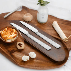 Styled flat lay of elegant dining setup featuring two silver textured dinner knives, white rolling pin, garlic cloves, small bowl with cheese, wooden board, and white bottle with fresh herbs on white background.