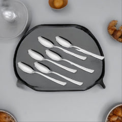 Six stainless steel teaspoons with textured handles arranged on dark hexagonal tray, surrounded by cashew nuts and glassware in styled setting.