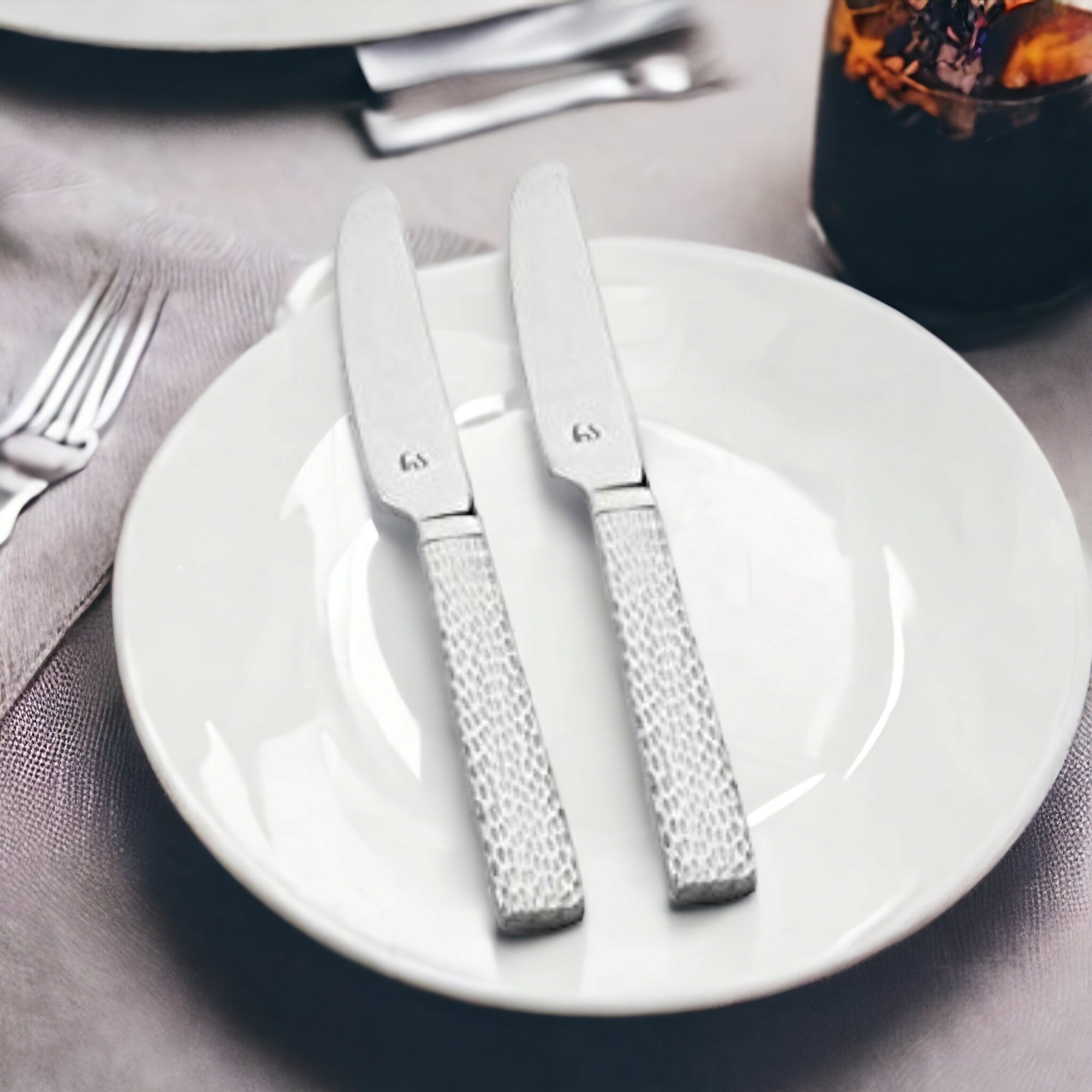 Two stainless steel dinner knives with textured silver handles placed on white plates beside fork and gray napkin in elegant table setting.
