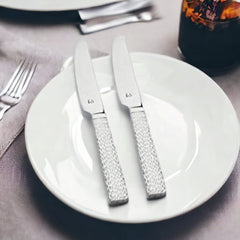 Two stainless steel dinner knives with textured silver handles placed on white plates beside fork and gray napkin in elegant table setting.