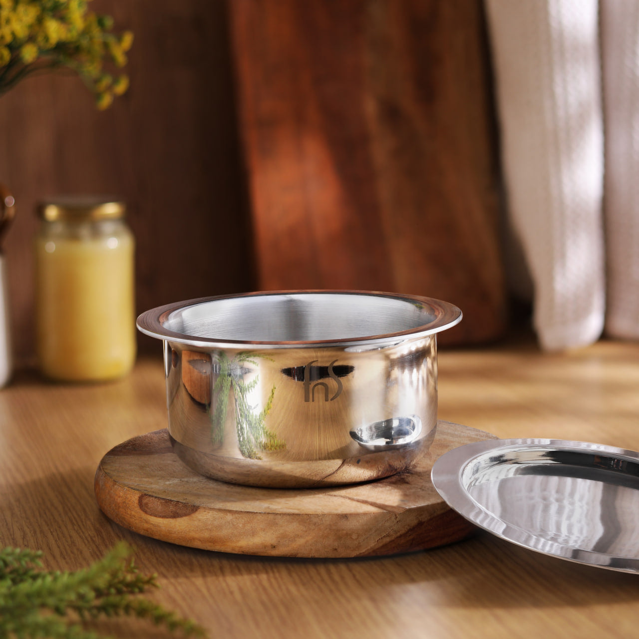 Stainless steel Tope with lid on wooden trivet, featuring metallic finish and minimalist design on warm-toned background.