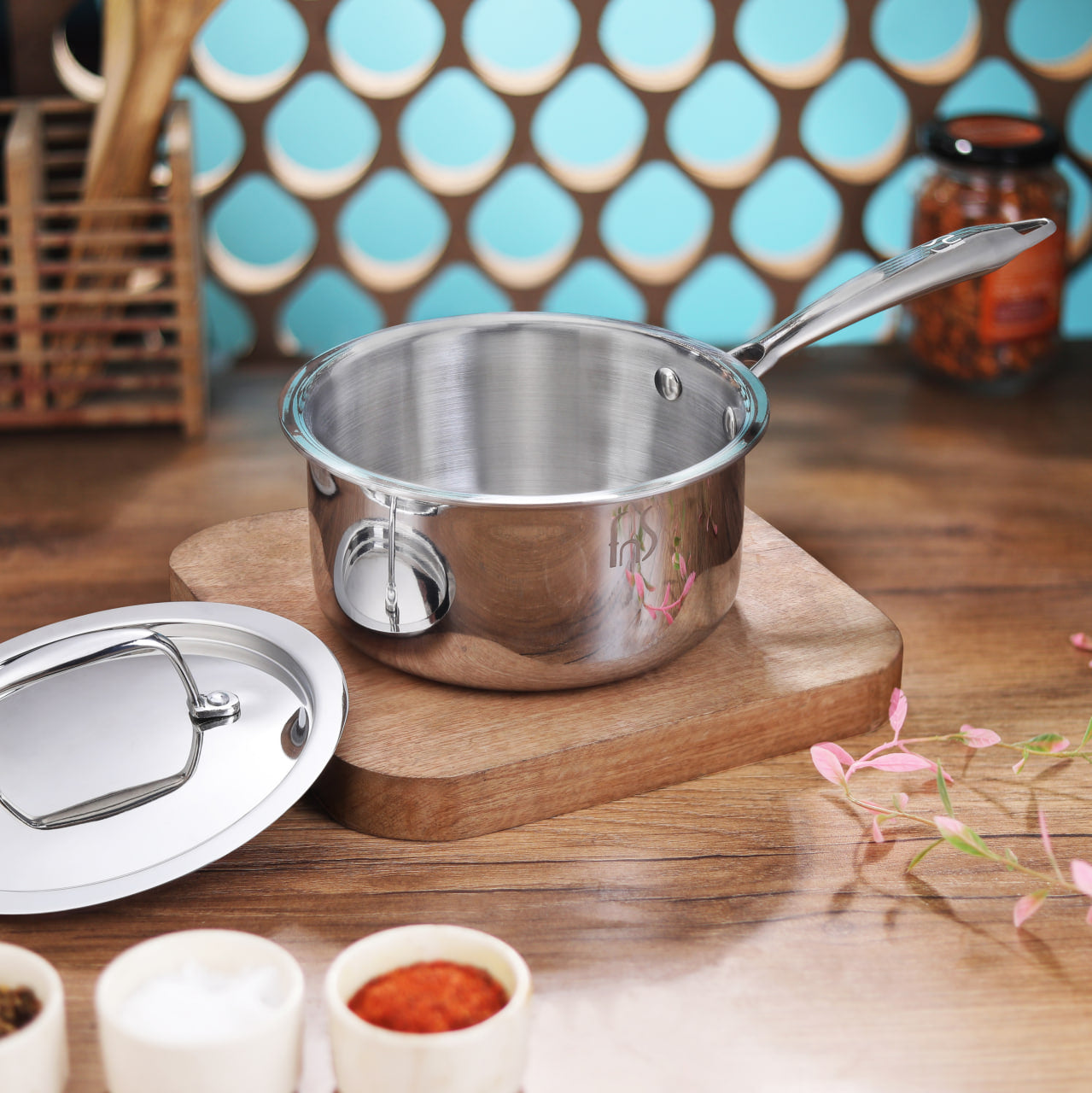 Stainless steel saucepan with handle and glass lid on wooden base, surrounded by cooking ingredients and decorative items.