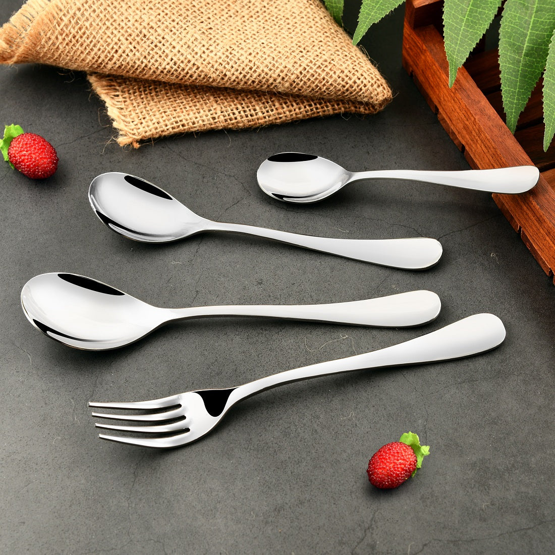 Four-piece silver flatware set laid out casually on a stone surface with strawberries and leaves.