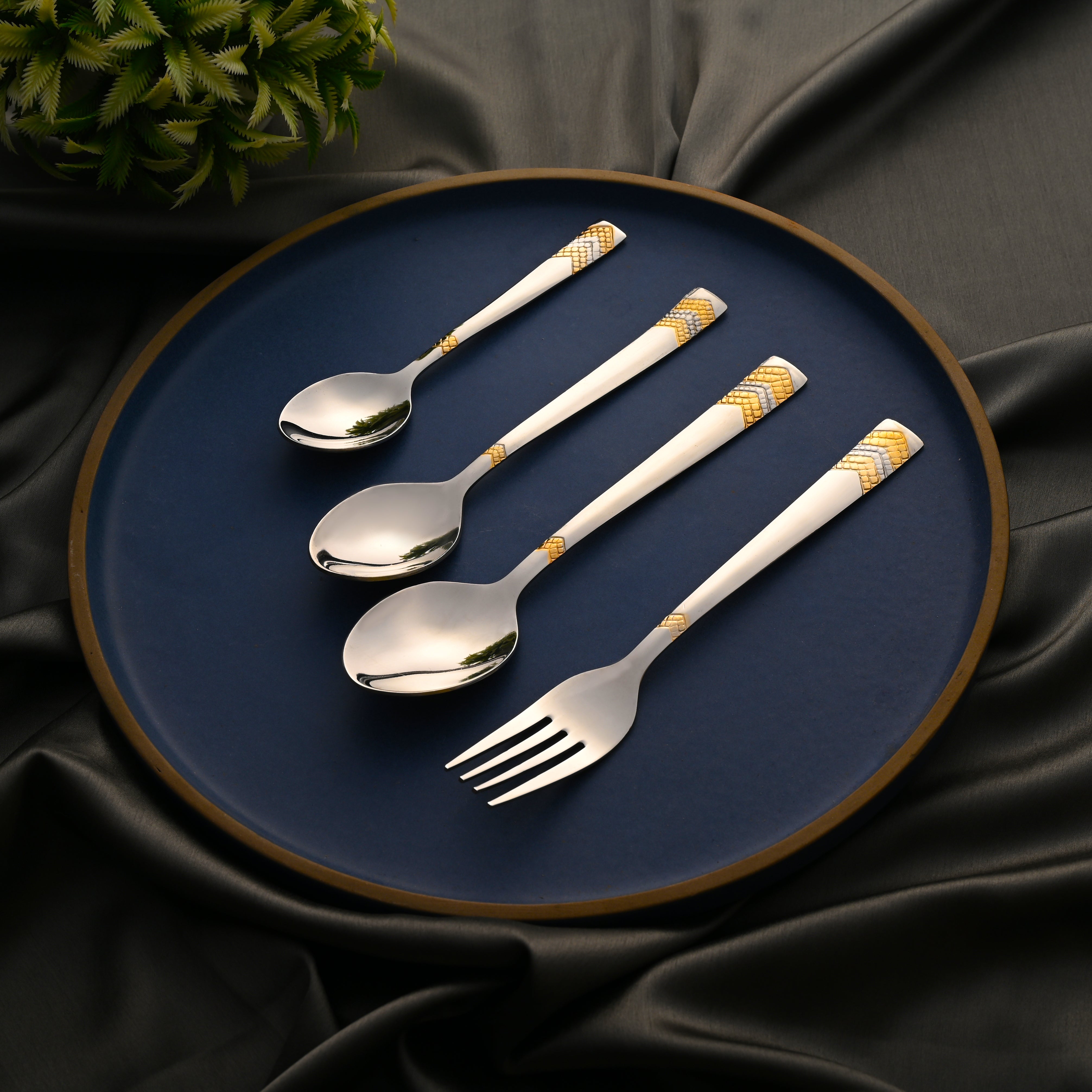Elegant gold-accented flatware set arranged on a blue plate with black satin and plant nearby.
