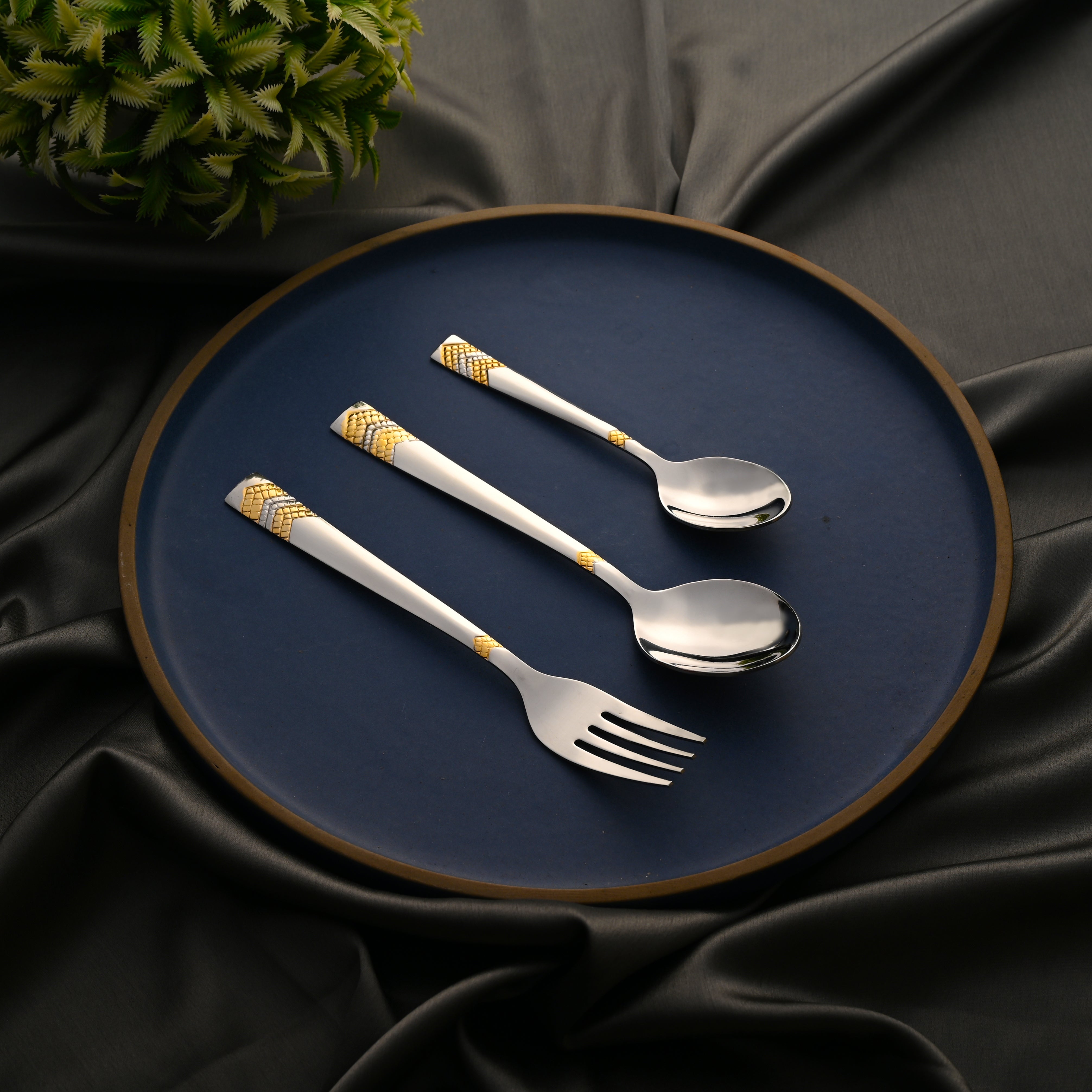 Cutlery trio with gold detailing arranged on a dark blue plate over smooth black satin fabric.