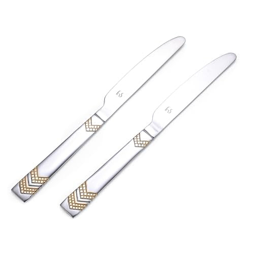 Two Raga dinner knives with gold chevron handles crossed beside layered cake with white frosting on elegant white serving plate.