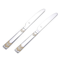 Two Raga dinner knives with gold chevron handles crossed beside layered cake with white frosting on elegant white serving plate.
