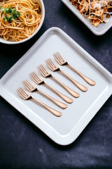 Set of six rose gold dinner forks displayed on a white tray, surrounded by pasta dishes, highlighting stylish and durable dining cutlery.