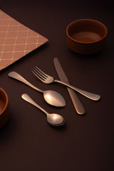 Styled flat lay showing rose gold dinner utensils on dark surface with decorative coral grid patterned napkin, terracotta bowl, and warm ambient lighting for elegant table setting.