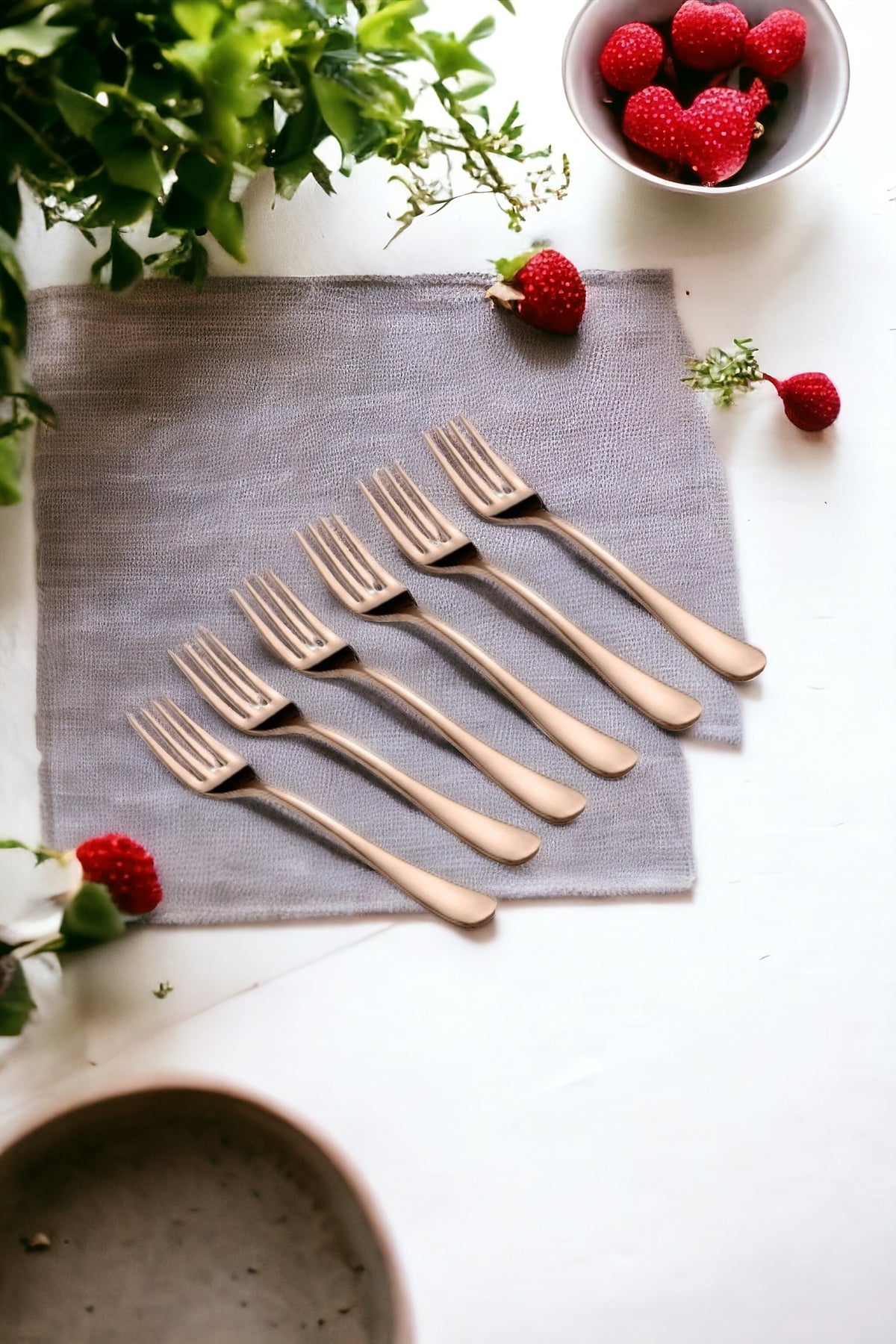 Six rose gold dinner forks arranged neatly on a grey cloth with strawberries and greenery around, emphasizing elegant tableware presentation.
