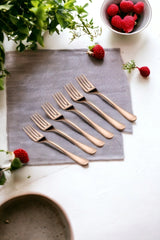 Six rose gold dinner forks arranged neatly on a grey cloth with strawberries and greenery around, emphasizing elegant tableware presentation.