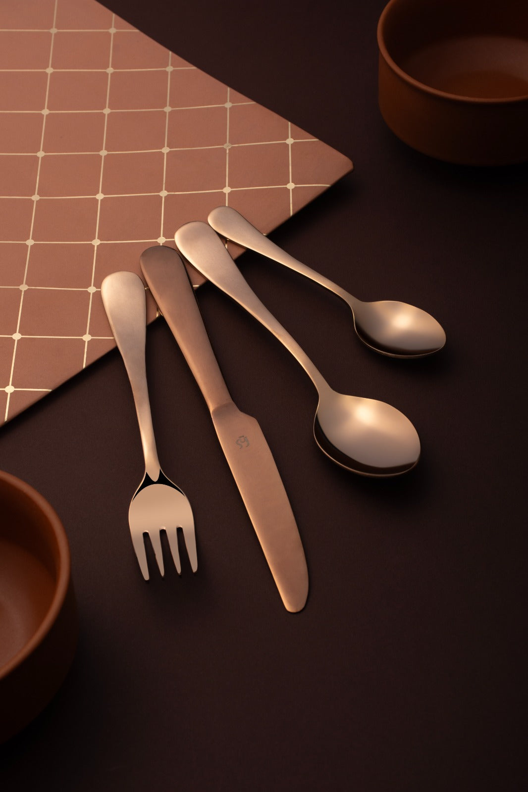 Overhead view of rose gold dinner fork, knife, and spoons arranged on coral geometric patterned napkin with terracotta bowl and dark surface, showcasing premium dining aesthetic.