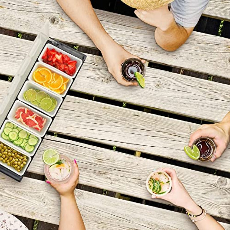 Overhead view of six-compartment condiment caddy filled with strawberries, citrus slices, grapefruit, cucumber, and olives on wooden deck table with people holding drinks