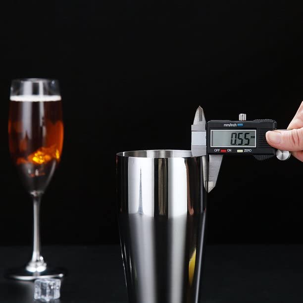 Stainless steel cocktail shaker being measured with digital caliper showing wall thickness with whiskey glass in background