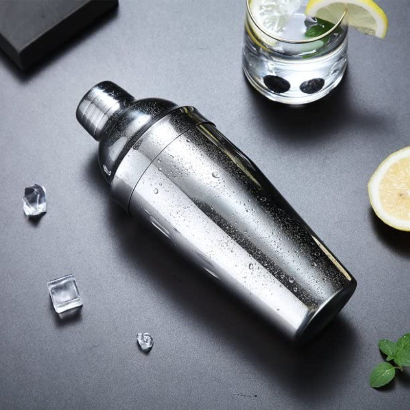 Stainless steel cocktail shaker with condensation on grey surface with ice cubes, lemon slices, mint, and gin and tonic glass