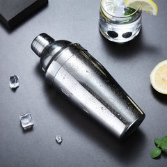 Stainless steel cocktail shaker with condensation on grey surface with ice cubes, lemon slices, mint, and gin and tonic glass
