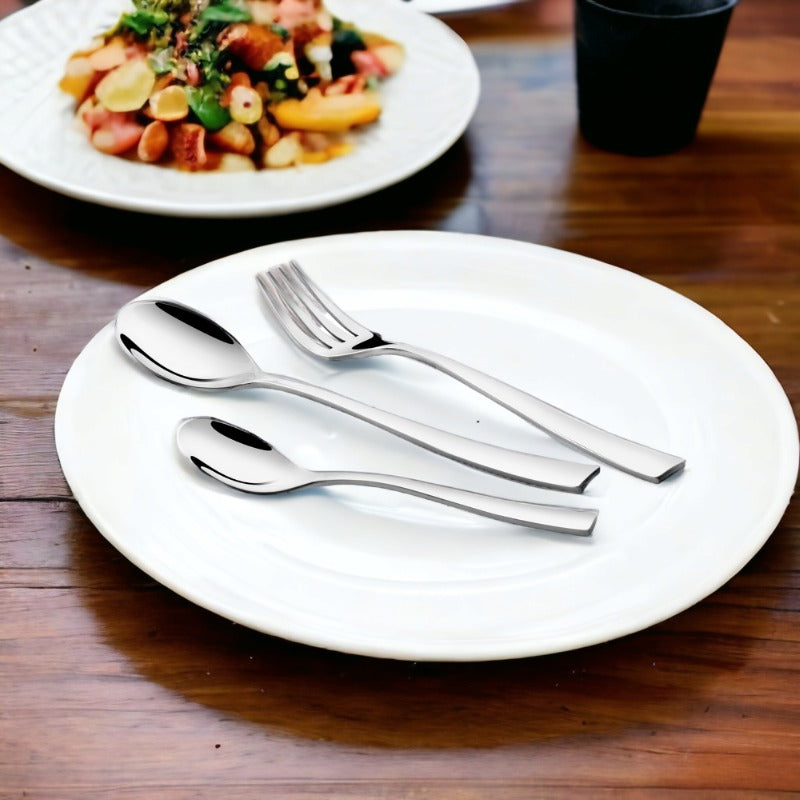 Stainless steel fork and spoons arranged on a white plate with a salad dish in background.