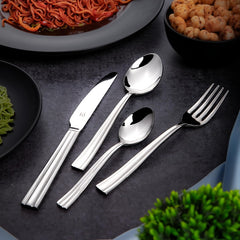 Shiny stainless steel FNS cutlery set placed on dark table with noodles, chickpeas, and greenery.