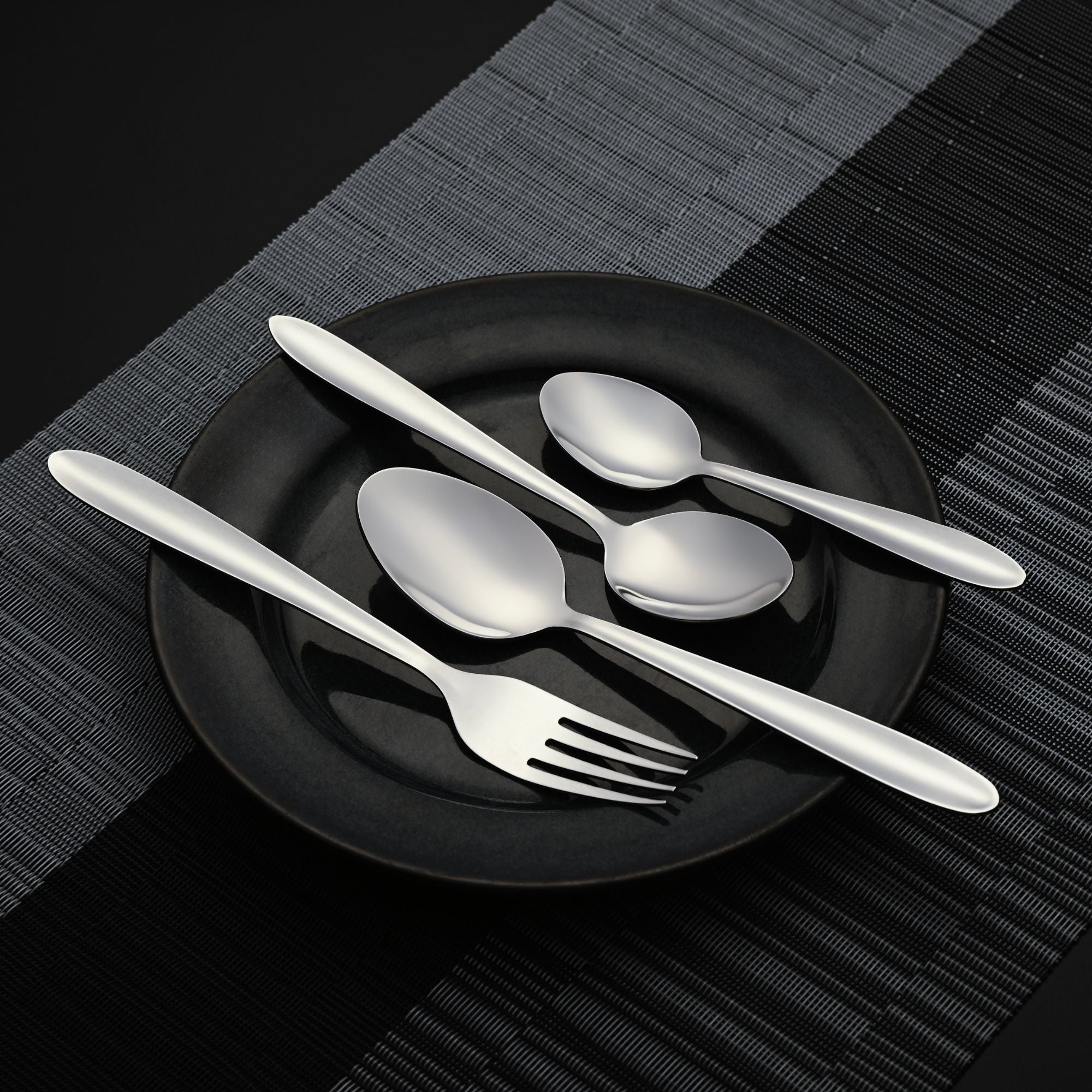 Four-piece stainless steel cutlery set arranged on black plate over dark striped placemat, minimalist styling.