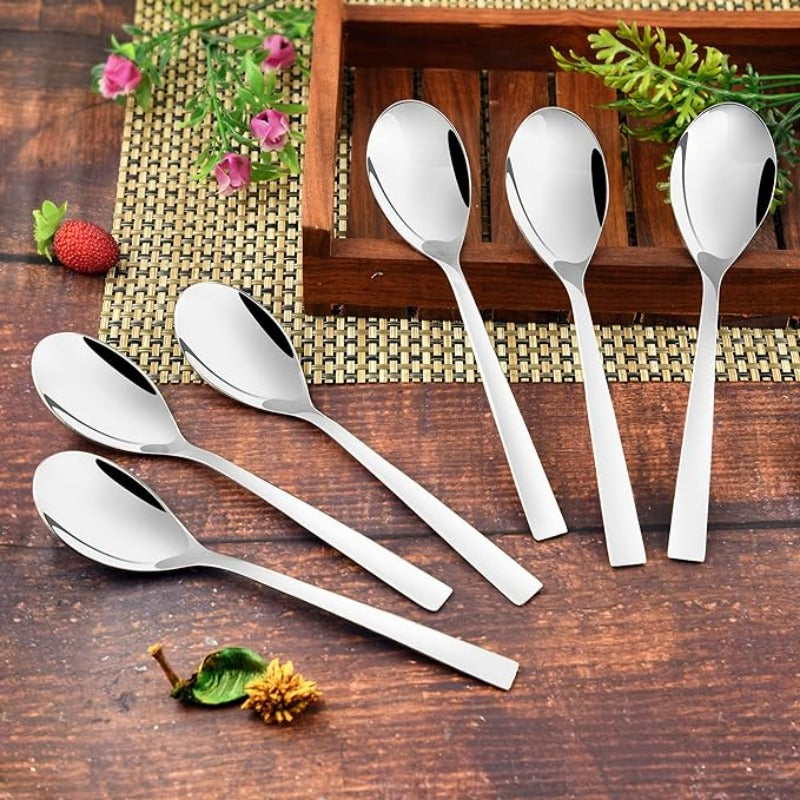 Six stainless steel dessert spoons arranged on wooden table with bamboo mat, wooden tray, pink flowers and strawberry